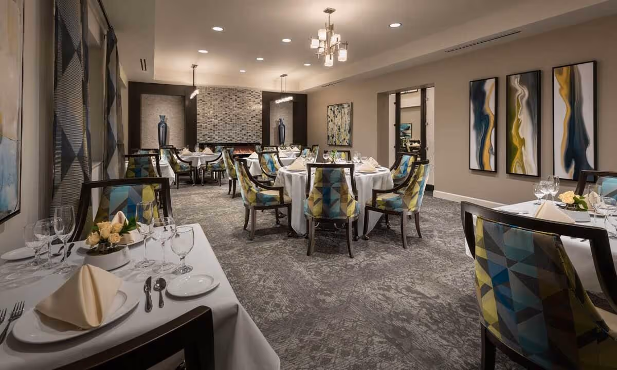 Elegant dining room with round tables covered in white tablecloths, set with napkins, plates, glasses, and silverware. Chairs have colorful geometric patterned upholstery. The room features modern lighting fixtures, abstract wall art, and a decorative fireplace with vases on either side.