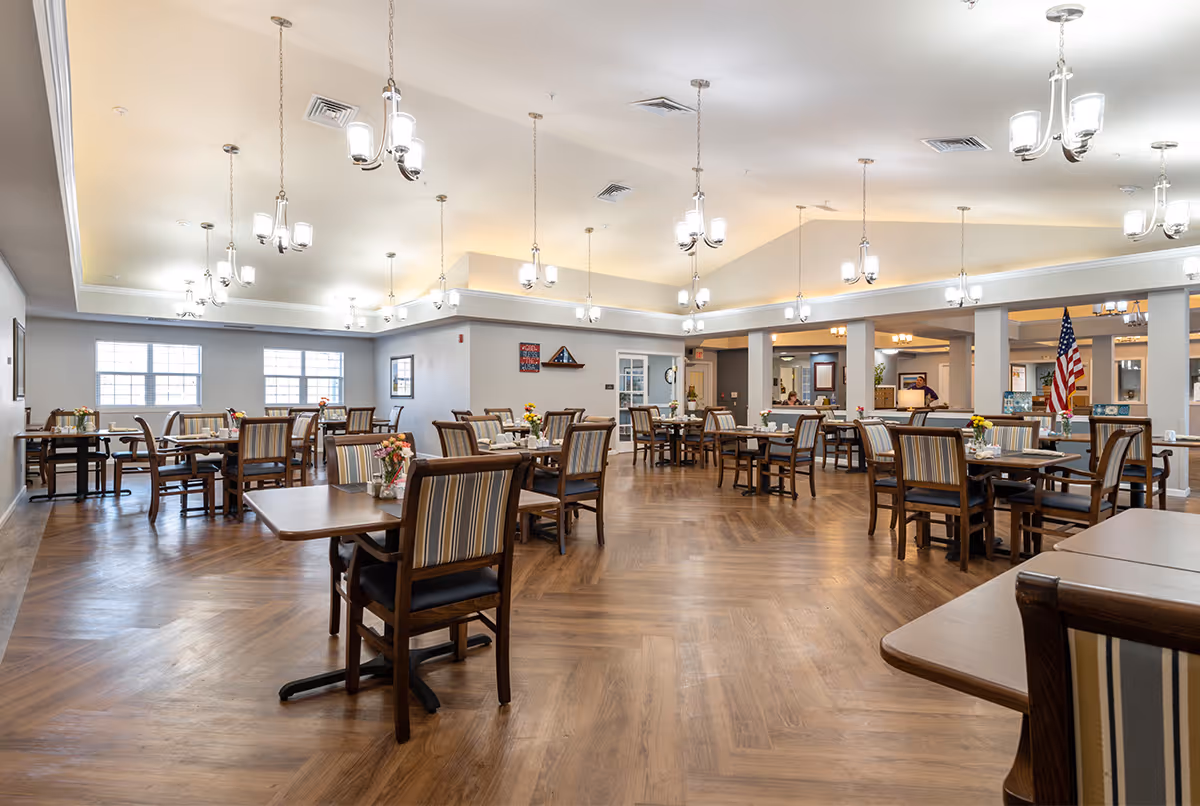 A spacious and well-lit dining room in a senior living facility with multiple wooden tables and chairs arranged neatly. The room features large windows, hanging light fixtures, and a wooden floor with a herringbone pattern. There are small flower arrangements on the tables and an American flag visible in the background.