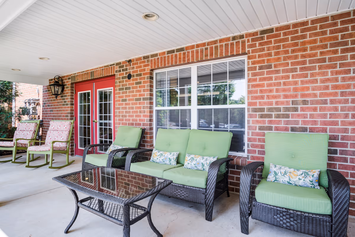 Covered outdoor patio area with brick walls, featuring green cushioned wicker chairs and a matching loveseat with floral pillows, a glass-top wicker coffee table, and two rocking chairs with floral cushions near red double doors.