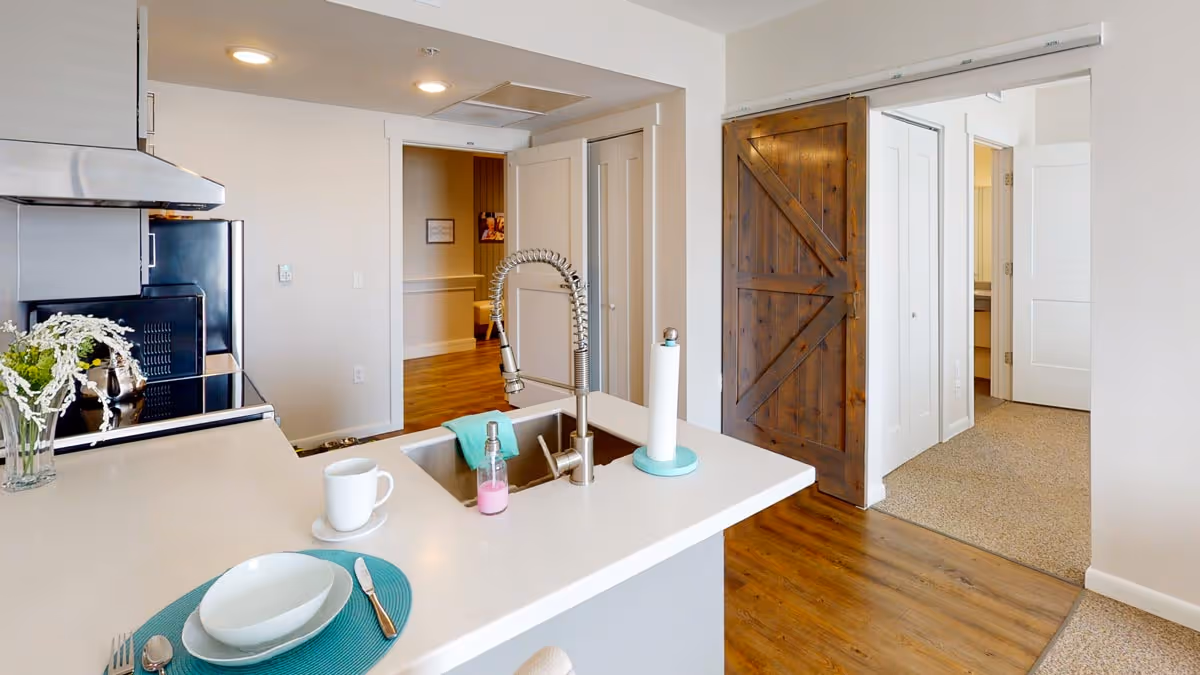 Modern kitchen area with a white countertop island featuring a sink with a flexible faucet, a soap dispenser, a paper towel holder, and a place setting with a plate, bowl, cup, and utensils on a teal placemat. Stainless steel appliances including a stove and microwave are visible on the left. The floor is wood, and there is a rustic sliding barn door on the right leading to another room with carpeted flooring.