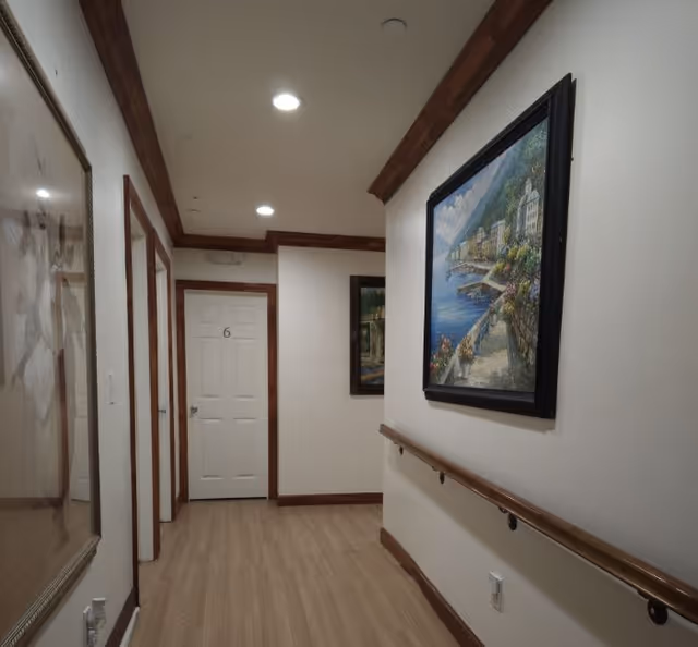 A well-lit hallway with wooden trim and handrails on the right wall. There are framed paintings hanging on the walls, and a white door labeled with the number 6 at the end of the hallway. The floor is light-colored wood, and the ceiling has recessed lighting.