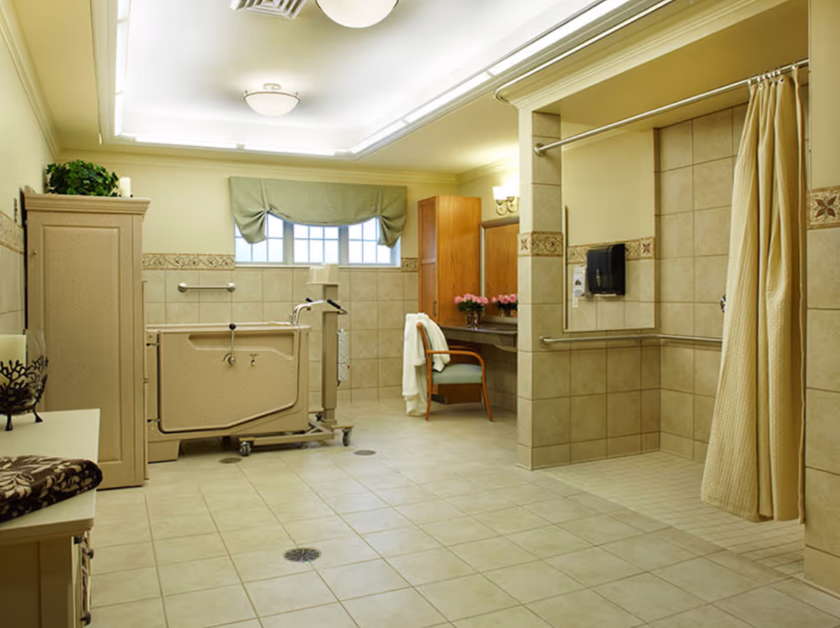 A spacious, well-lit bathroom with beige tiled floors and walls. The room features a walk-in shower with a yellow curtain, grab bars, and a wall-mounted soap dispenser. There is a large bathtub with a mechanical lift for assisted bathing. A wooden cabinet and a vanity area with a chair and flowers are visible in the background. A window with a green valance allows natural light into the room.