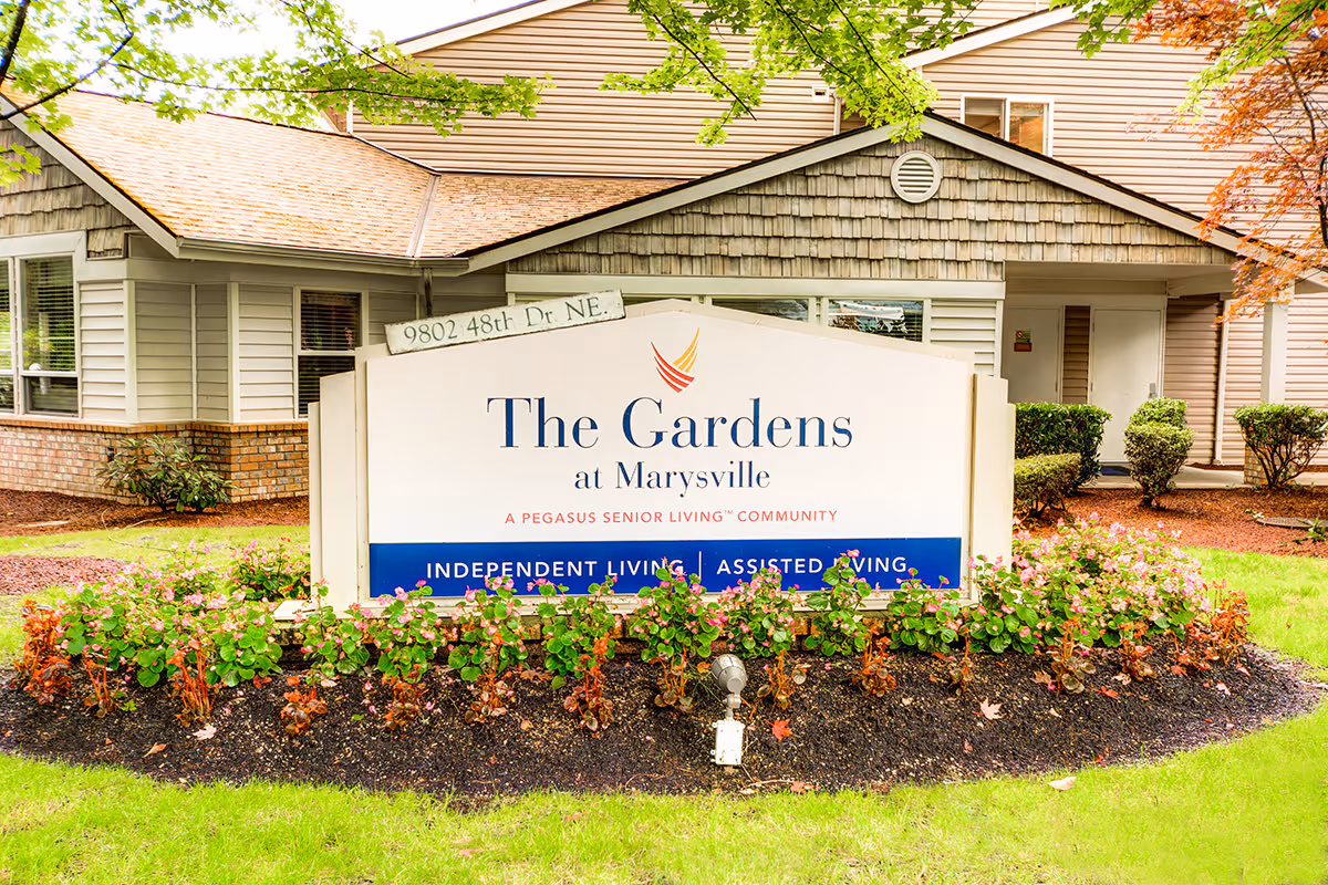 Outdoor view of the entrance sign for The Gardens at Marysville, a Pegasus Senior Living community offering independent living and assisted living. The sign is surrounded by flower beds and greenery, with part of the building visible in the background.