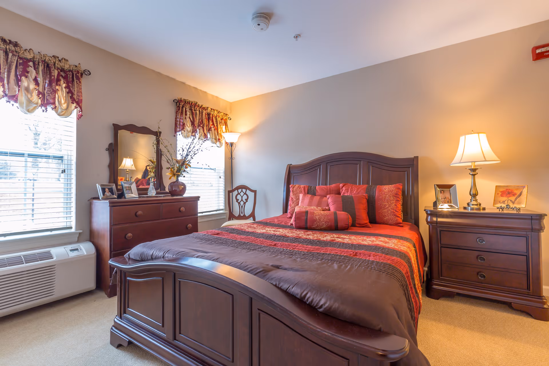 A warmly lit bedroom featuring a large wooden bed with a red and brown patterned comforter and multiple pillows. To the right of the bed is a wooden nightstand with a lamp and framed pictures. On the left side of the room, there is a wooden dresser with a mirror, decorative items, and framed photos. Two windows with red and gold valances allow natural light into the room. A wooden chair and a floor lamp are positioned near the windows.