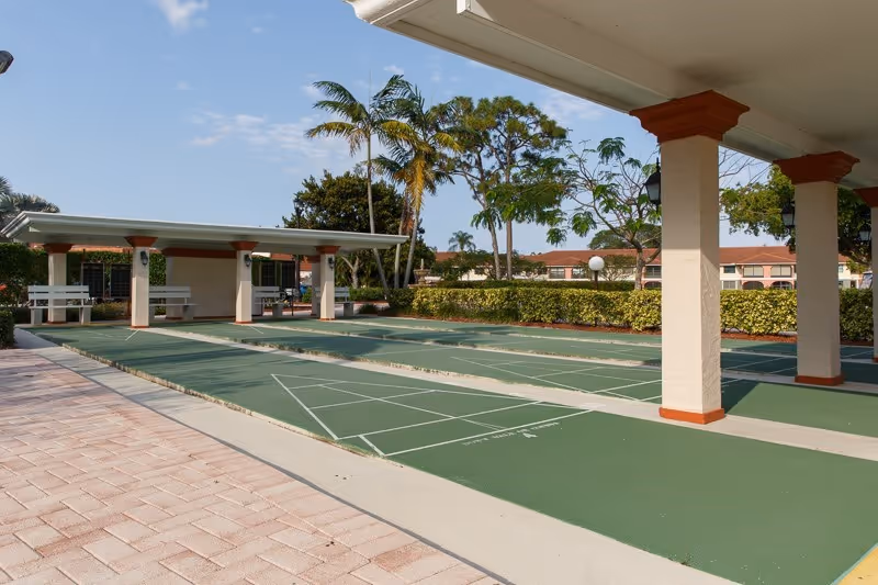 Outdoor shuffleboard courts with green playing surfaces and white markings, covered seating areas with benches, surrounded by palm trees and shrubbery under a clear blue sky.