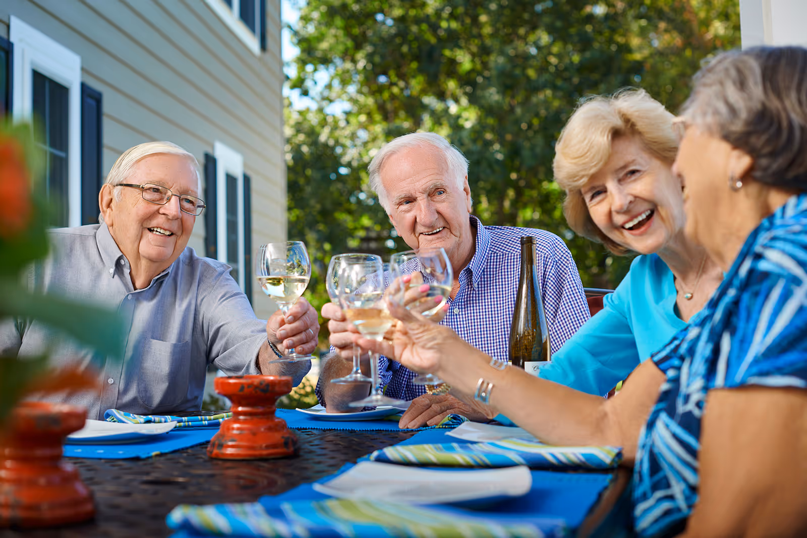 Four elderly people sitting outdoors around a table, smiling and raising glasses of white wine in a toast. The setting is a patio area with greenery and a building with windows in the background.