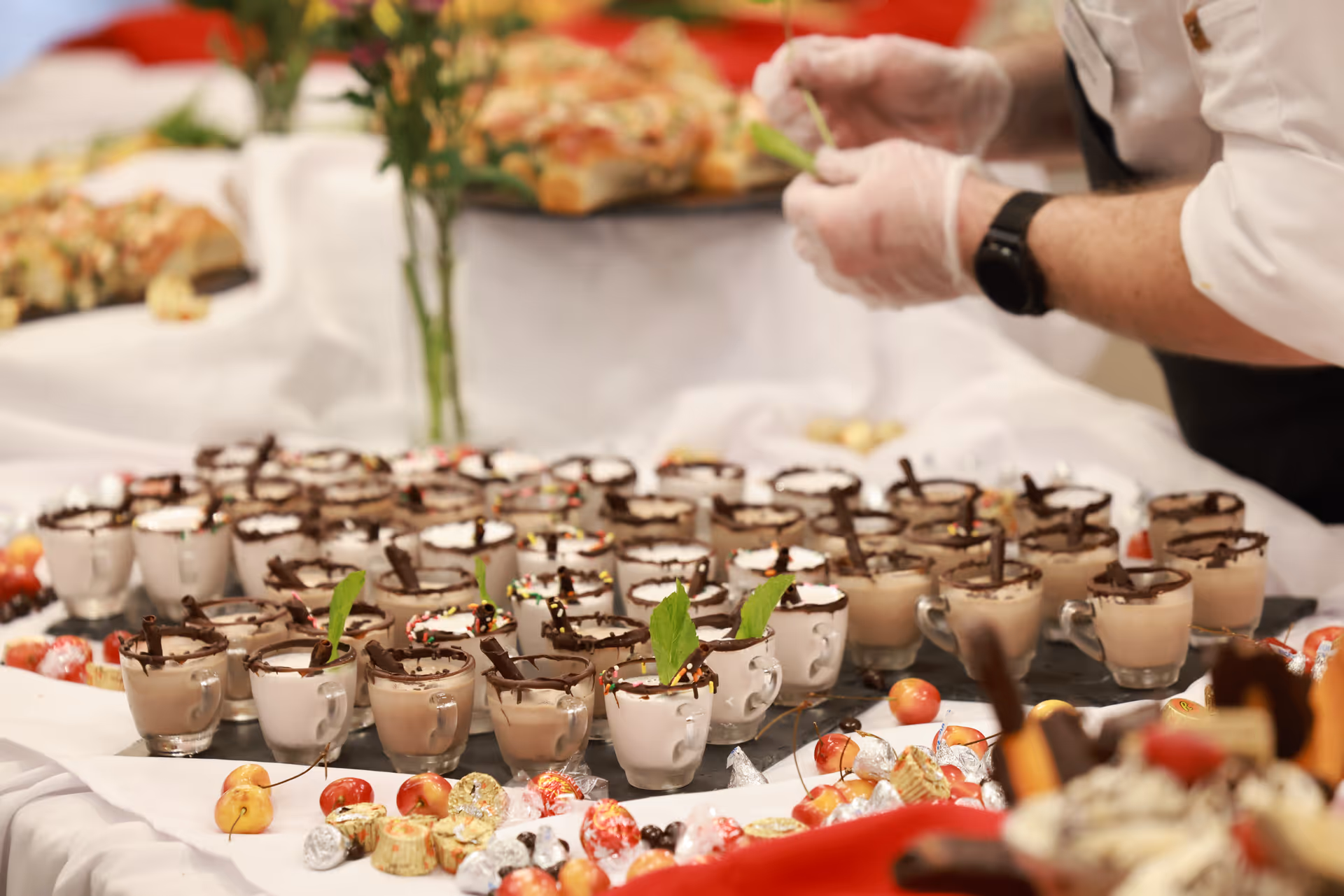 A table filled with multiple small glass cups of chocolate mousse or dessert, each garnished with chocolate sticks and some with mint leaves. A person wearing gloves is seen preparing or arranging the desserts. The table is decorated with small fruits and wrapped candies.