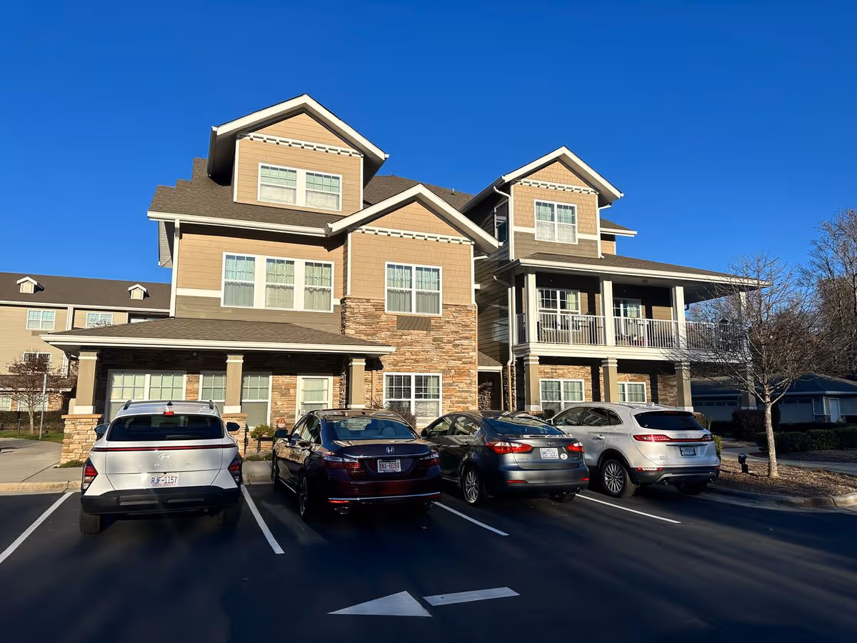 Front exterior of a multi-story beige senior living building with a covered porch and several cars parked in front.