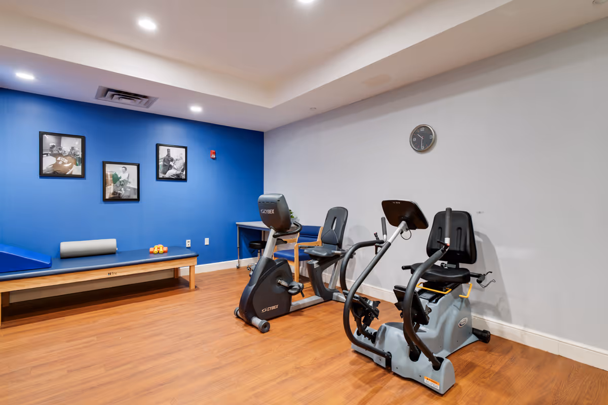 A small exercise room with wooden flooring, featuring two stationary exercise bikes and a padded bench against a blue wall. The blue wall has three black and white framed photographs, and there is a clock on the adjacent white wall. A small desk with a chair is also visible in the corner.