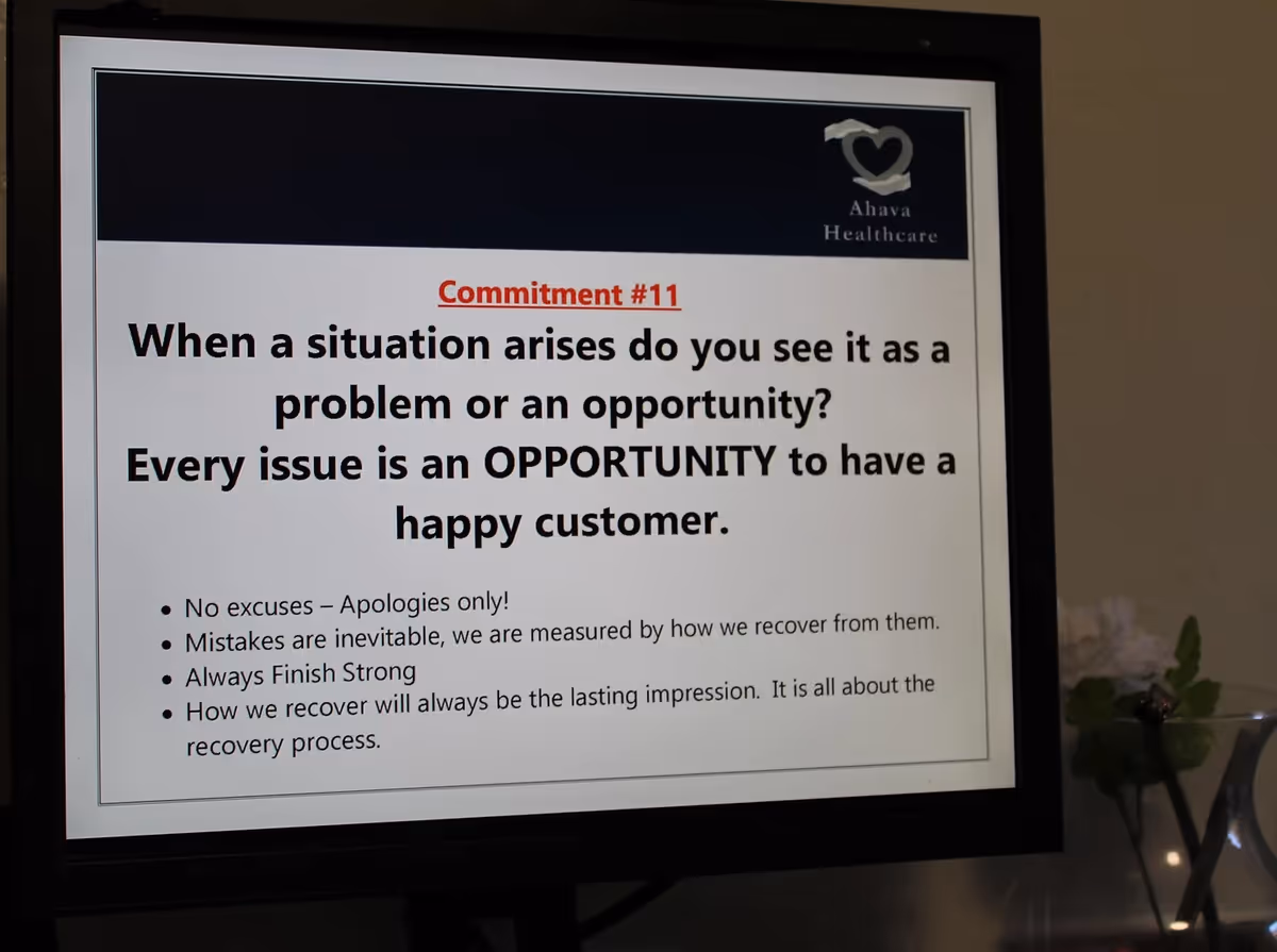 A computer monitor displaying a slide titled Commitment #11 from Ahava Healthcare. The slide reads: When a situation arises do you see it as a problem or an opportunity? Every issue is an OPPORTUNITY to have a happy customer. Below are bullet points: No excuses – Apologies only! Mistakes are inevitable, we are measured by how we recover from them. Always Finish Strong. How we recover will always be the lasting impression. It is all about the recovery process.