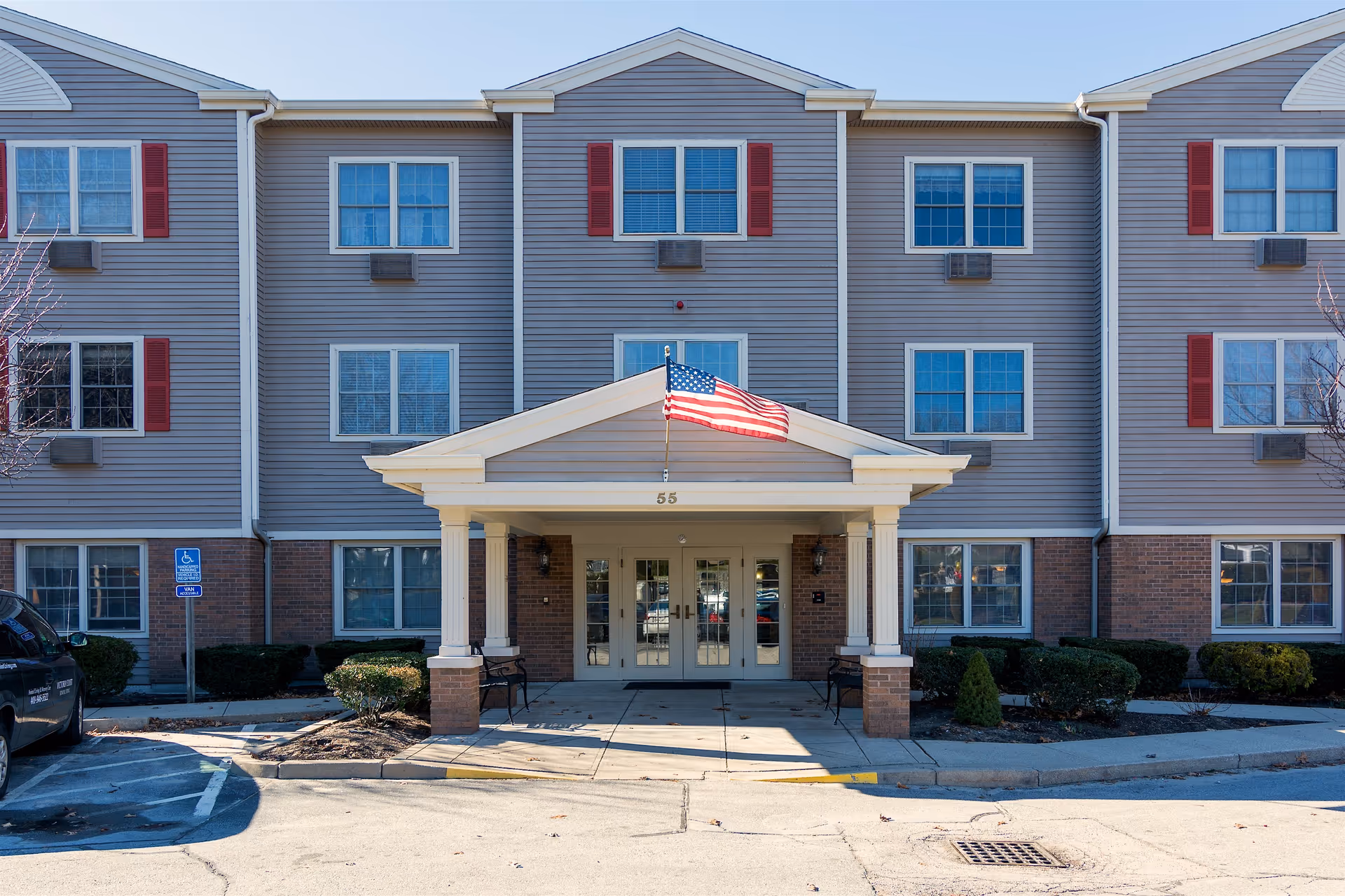 Exterior front entrance of a three-story building with gray siding and red shutters. The entrance has a covered porch with white columns and an American flag mounted above the door. There are benches on either side of the entrance and a handicapped parking sign visible on the left side.