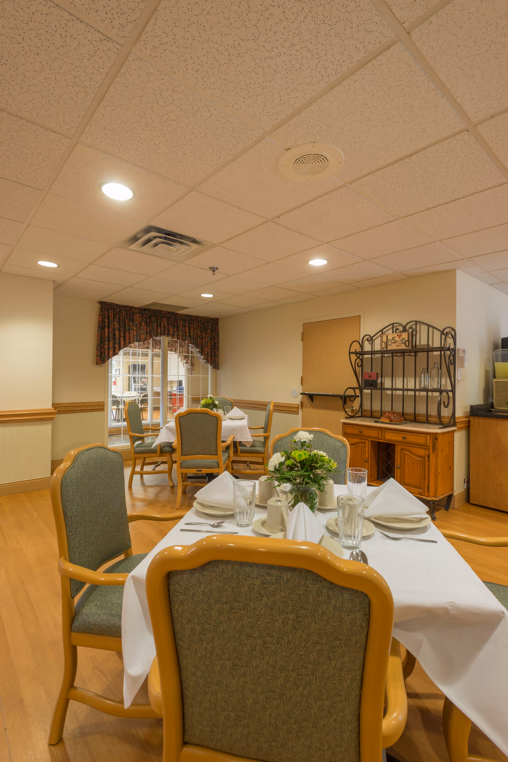 Dining area with set tables and chairs, floral centerpieces, and a sideboard in a senior facility.