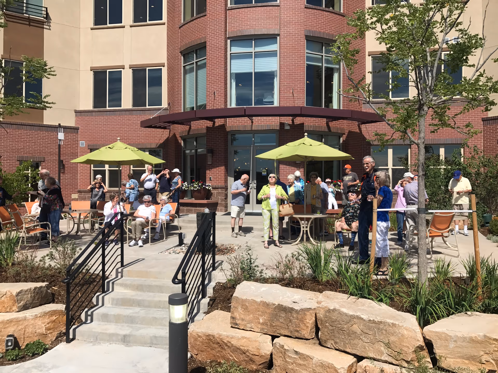 Outdoor patio area at a senior living facility with several elderly people sitting and standing around tables with green umbrellas. The building has a brick and beige exterior with large windows. There are plants and rocks in the foreground and a set of stairs leading up to the patio.