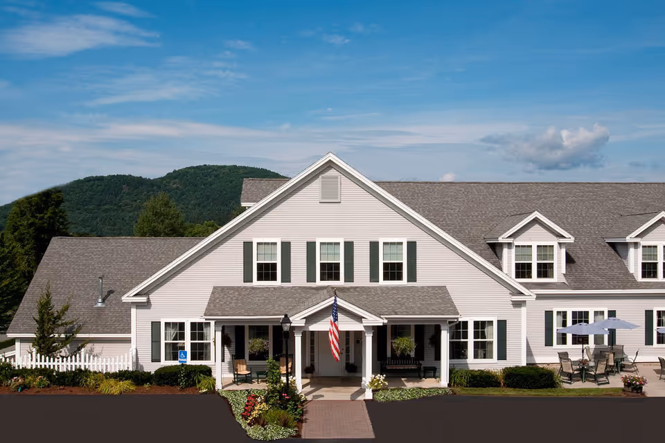 Front exterior of a white senior living building with a porch, American flag, patio seating, and tree-covered hills in the background.