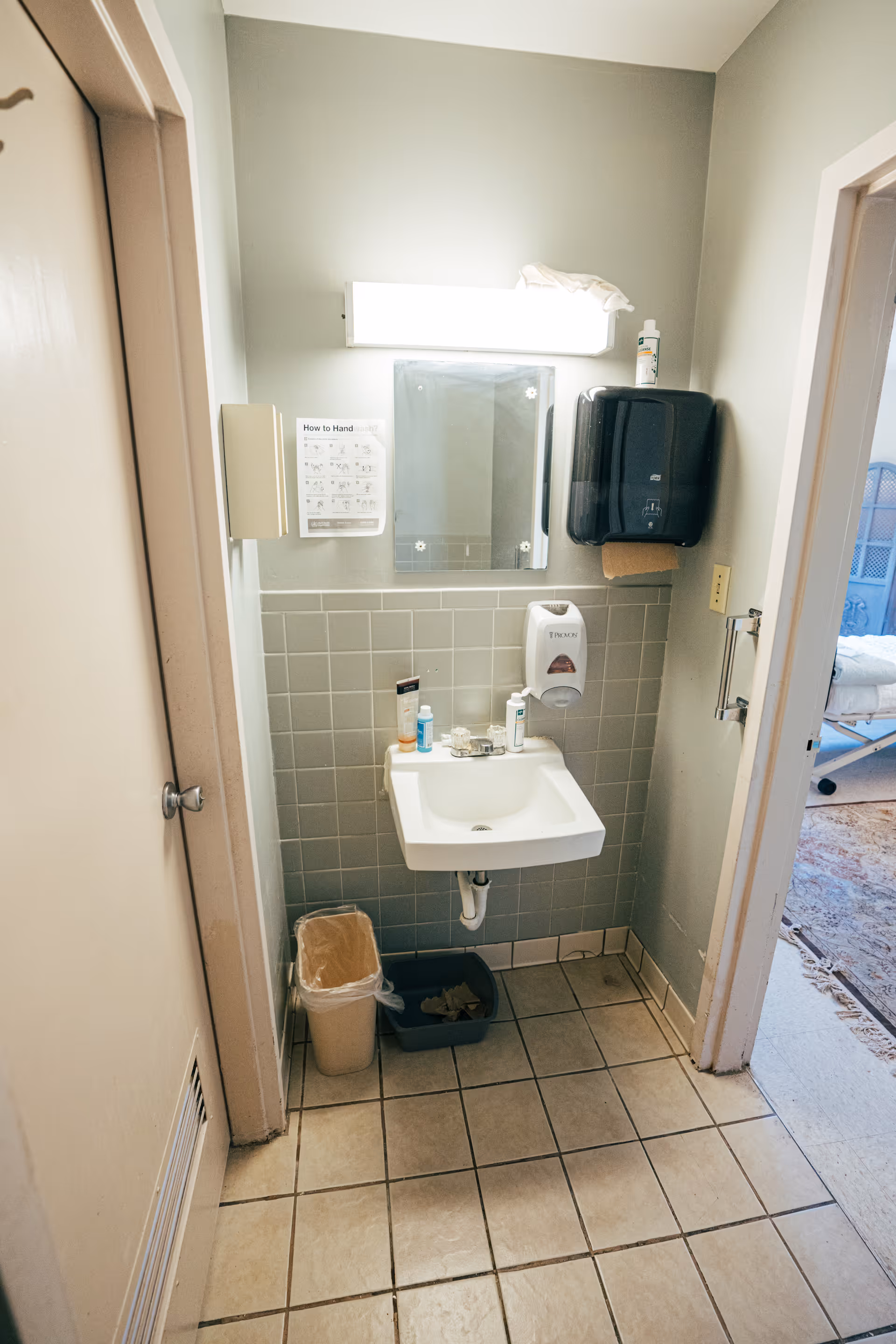 Small bathroom area with a wall-mounted sink, a mirror above it, a soap dispenser, a paper towel dispenser, and a trash can below. There is a tiled wall and floor, and a sign on the wall with handwashing instructions. A doorway leads to another room with a bed visible.