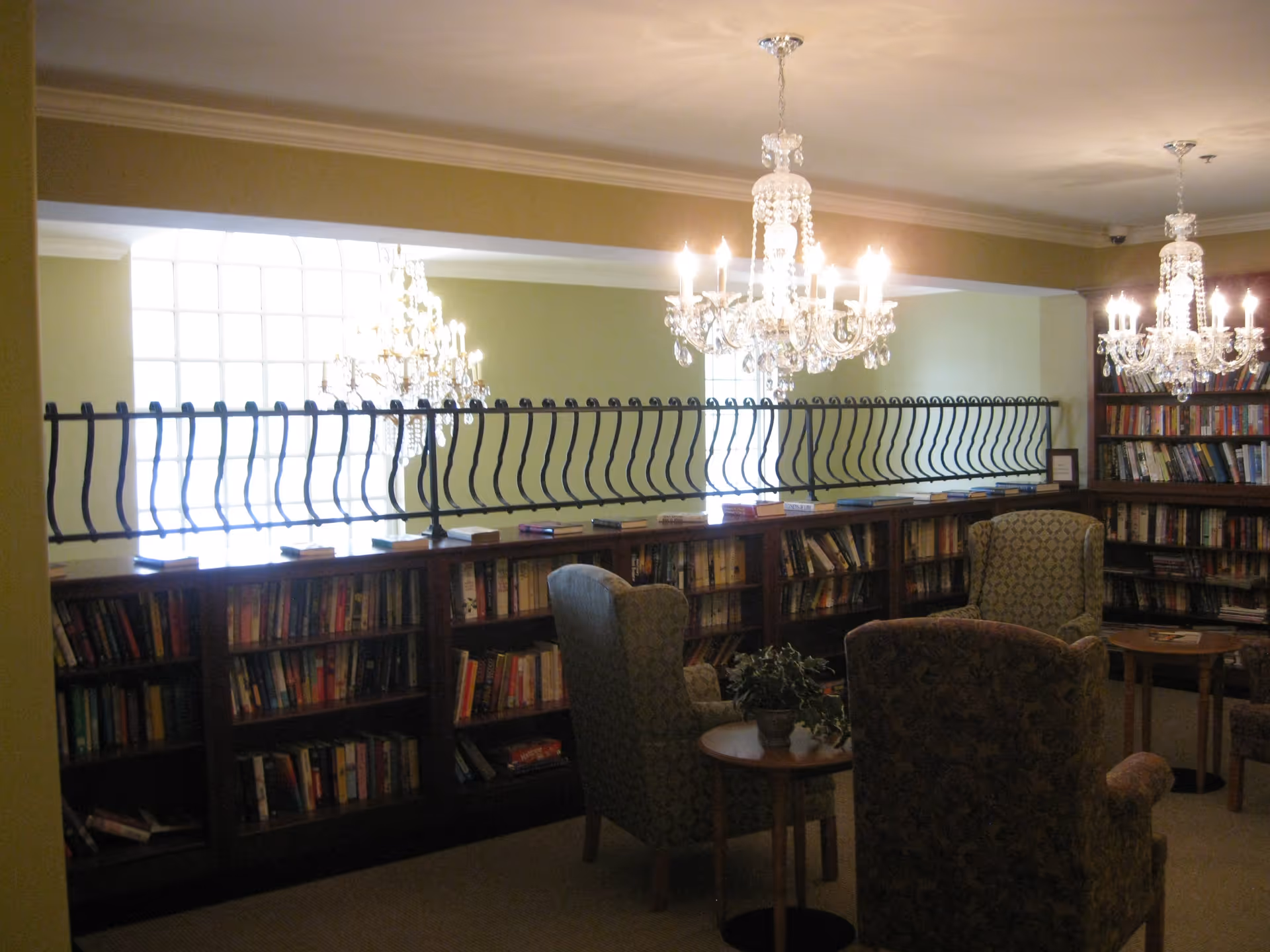 Cozy senior living library/reading room with bookshelves, upholstered armchairs, small tables, and crystal chandeliers.