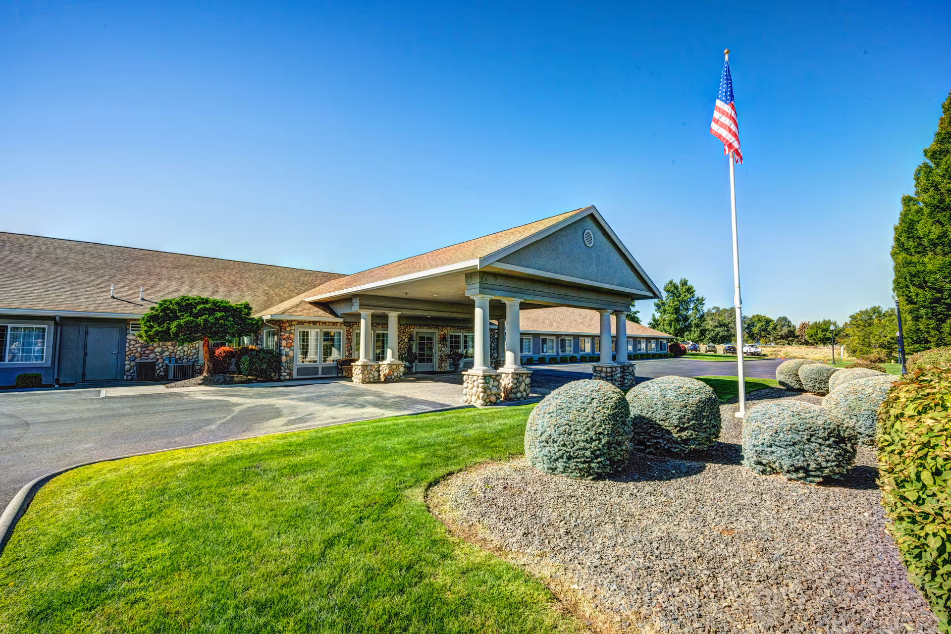 Exterior view of Callaway Gardens Alzheimer's Special Care facility showing a single-story building with a covered entrance supported by stone pillars, a well-maintained lawn, neatly trimmed bushes, and an American flag on a flagpole under a clear blue sky.