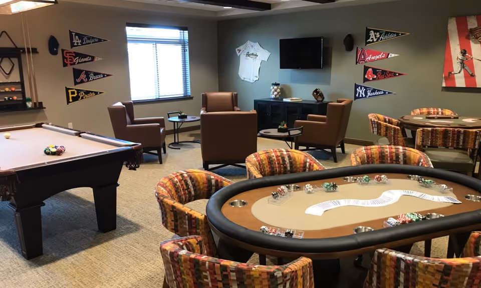 A recreational room with a poker table in the foreground surrounded by colorful patterned chairs. Behind it, there is a pool table with billiard balls arranged on it. The room has several brown armchairs and small round tables near a window with blinds. The walls are decorated with various baseball team pennants and a framed baseball jersey, along with a flat-screen TV mounted on the wall.