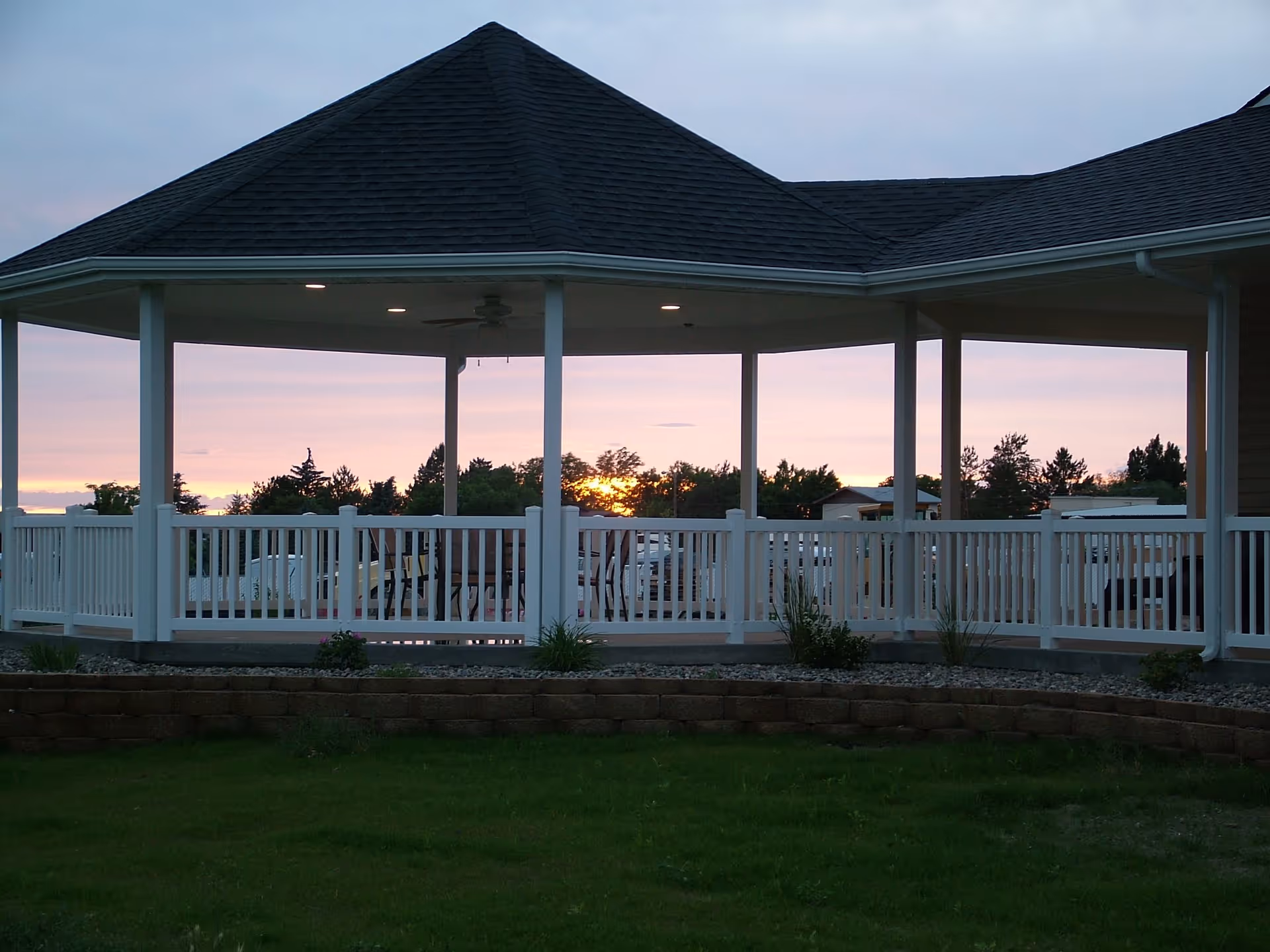 A covered outdoor gazebo with white railings and a dark shingled roof, set against a sunset sky with trees and buildings in the background. The gazebo is surrounded by a green lawn and a low stone retaining wall.