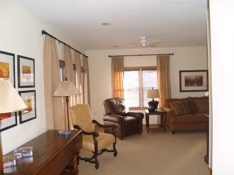 A cozy living room with beige walls and carpet, featuring a brown leather armchair, a patterned fabric armchair, and a brown sofa with decorative pillows. There are two windows with beige curtains and wooden blinds, a wooden side table with a lamp, a wooden table with a lamp and some boxes on it, and framed artwork on the walls.