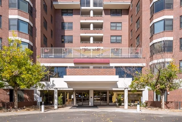 Front exterior view of a multi-story brick building with large windows and balconies. The entrance has a covered drop-off area supported by white columns, with trees and landscaping on either side.