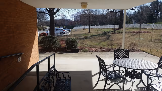 Covered patio with a metal table and chairs and a bench overlooking a grassy yard, trees, and parked cars.