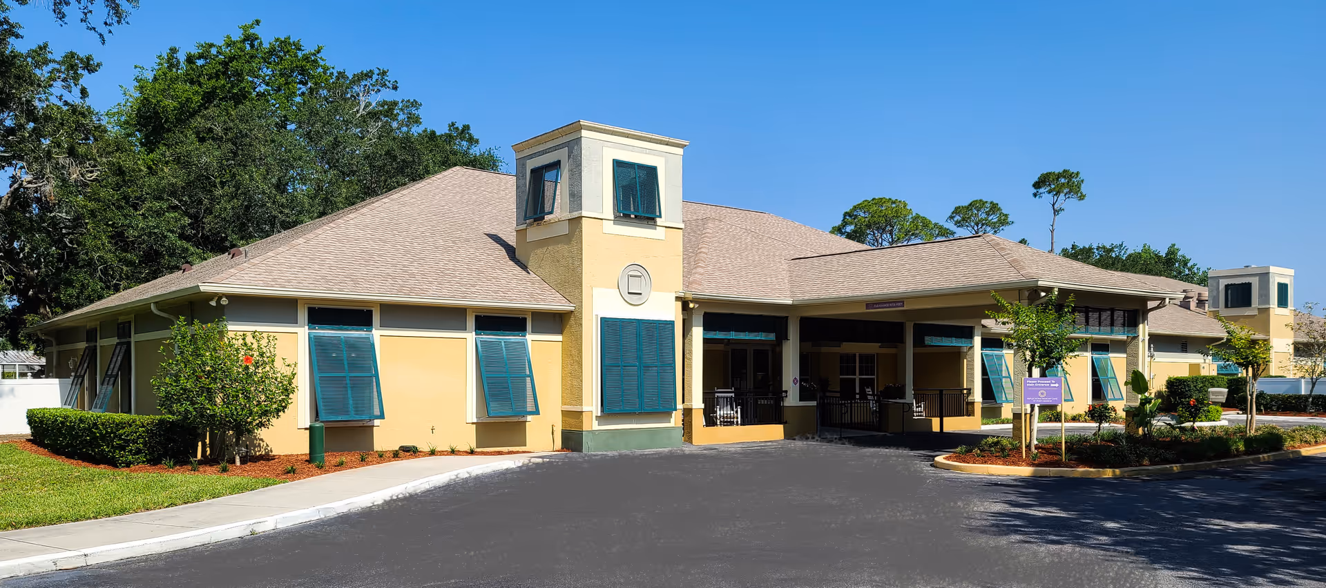 Exterior view of a single-story senior living facility building with beige walls, green window shutters, and a tan roof. The building has a covered entrance with a driveway in front, surrounded by landscaped greenery and trees under a clear blue sky.