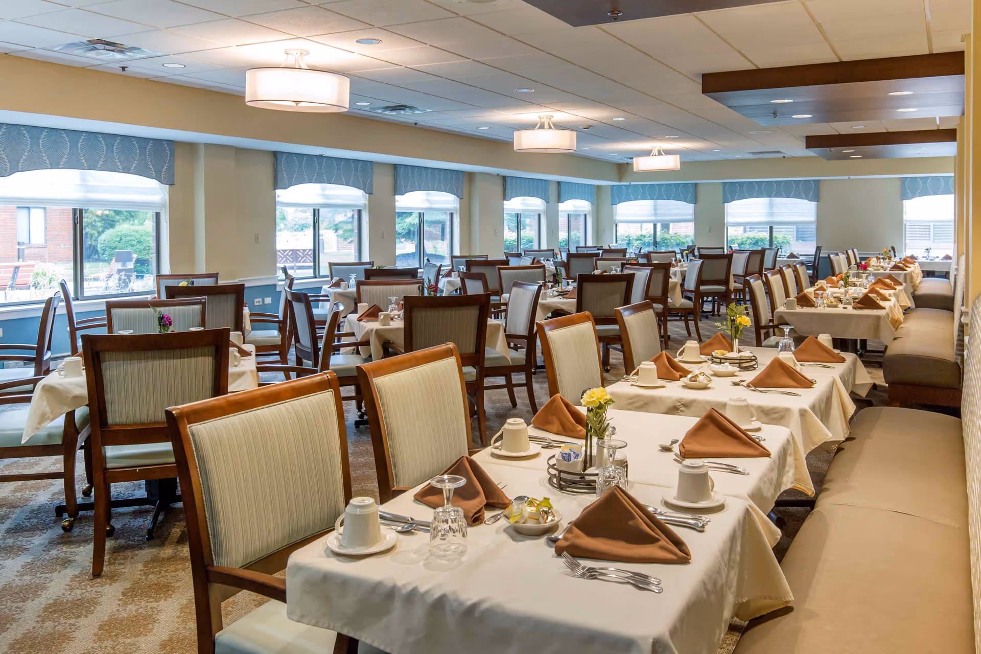 A spacious dining room with multiple tables set for a meal, featuring white tablecloths, brown napkins, cups, and silverware. The room has large windows with blue valances allowing natural light to enter, and cushioned chairs arranged neatly around the tables.