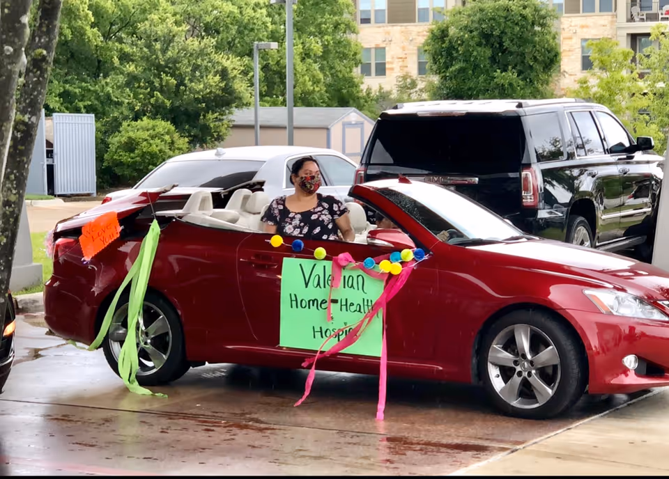 A red convertible decorated with colorful signs, streamers, and pom-poms parked outside with a masked woman seated inside.