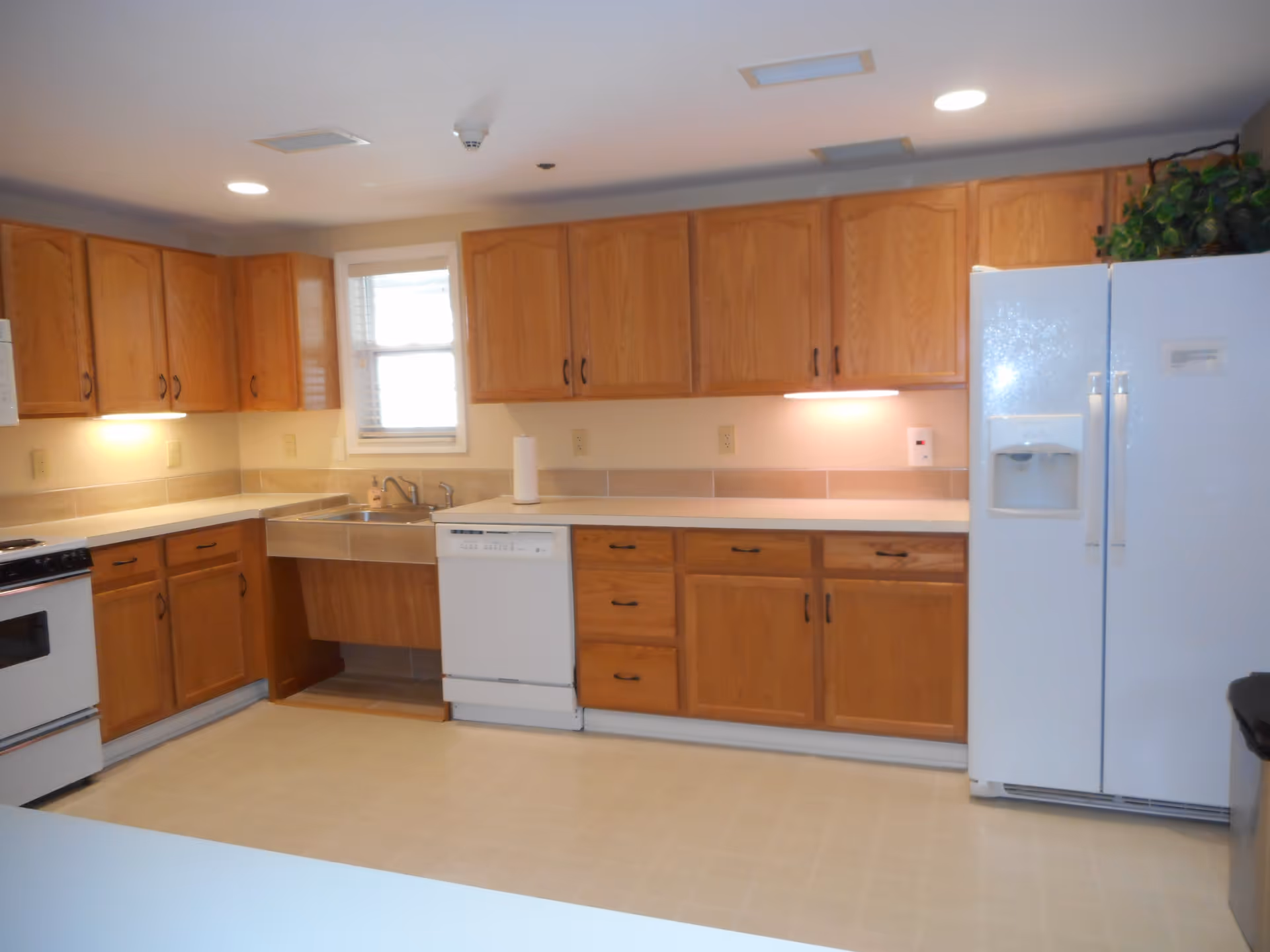 A clean kitchen with wooden cabinets, a white refrigerator with a water dispenser, a dishwasher, a stove, a sink under a small window, and beige tiled backsplash and floor.