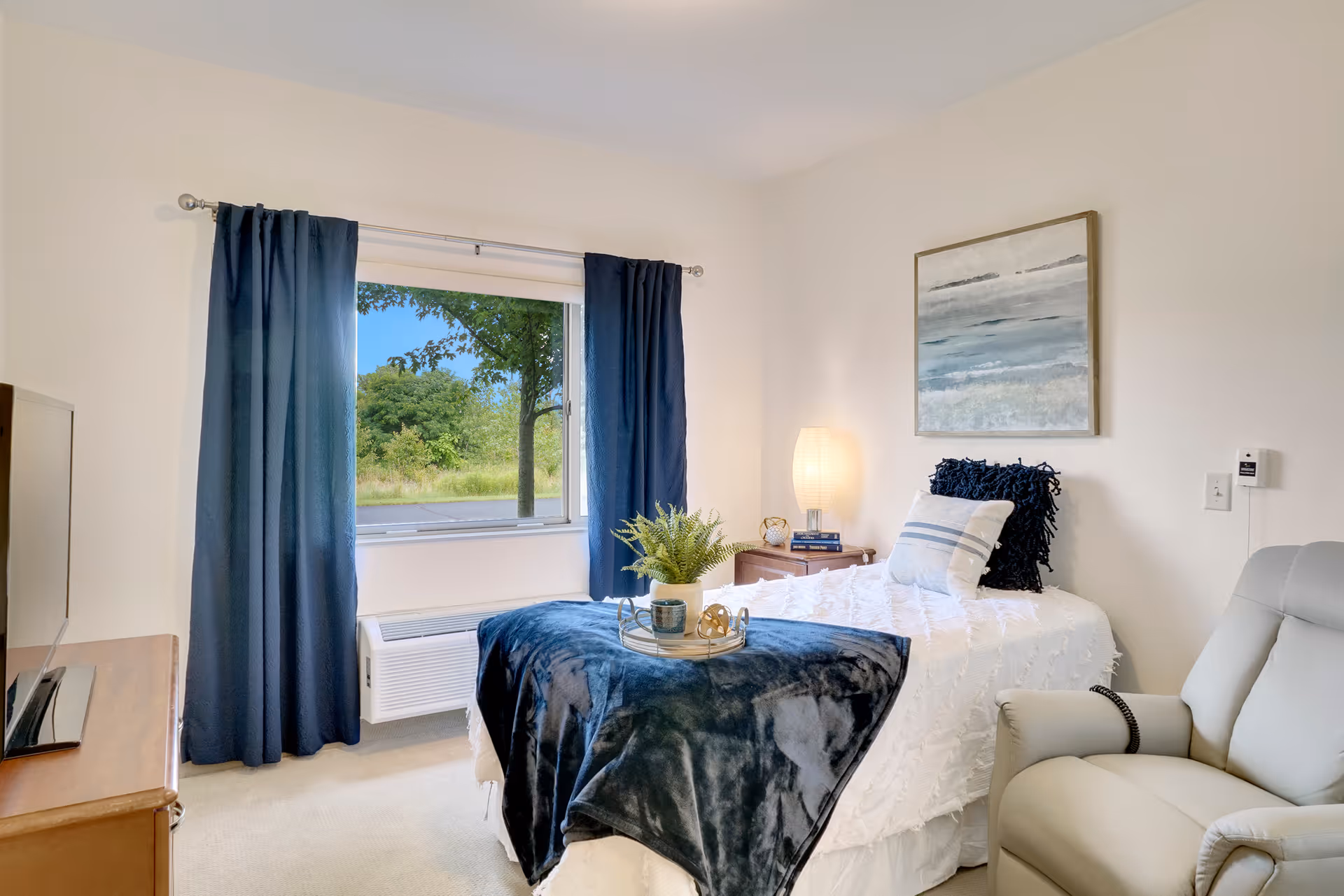 A bright and cozy bedroom with a single bed covered in white bedding and a dark blue throw blanket. There are two pillows on the bed, one white with blue stripes and one dark blue. A small wooden nightstand with a lamp and decorative items is beside the bed. A large window with dark blue curtains shows a view of green trees outside. A comfortable beige recliner chair is positioned near the bed, and a wooden dresser with a TV is visible on the left side of the room. A framed abstract painting hangs on the wall above the bed.