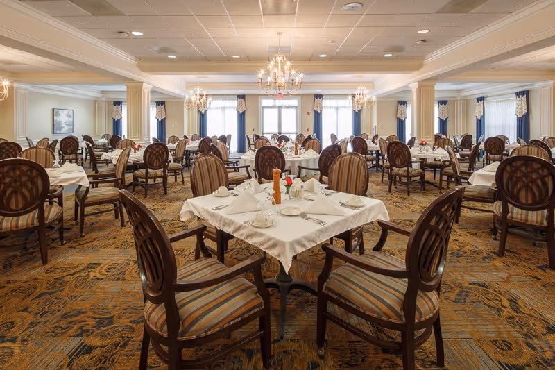 A spacious and elegant dining room with multiple tables covered in white tablecloths, each set with cups, plates, napkins, and silverware. The room features striped upholstered chairs, chandeliers hanging from the ceiling, large windows with curtains, and a patterned carpet.