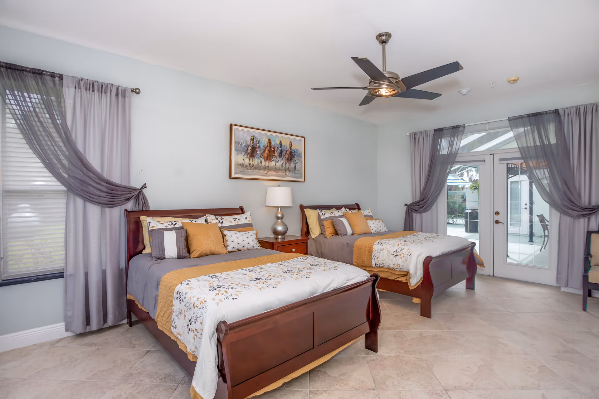 A bright bedroom with two wooden beds, each made with gray, white, and mustard yellow bedding and multiple pillows. Between the beds is a wooden nightstand with a silver lamp. Above the nightstand hangs a painting of horses racing. The room has light blue walls, beige tiled floor, a ceiling fan with lights, and large windows with sheer purple curtains. French doors lead to an outdoor patio area.
