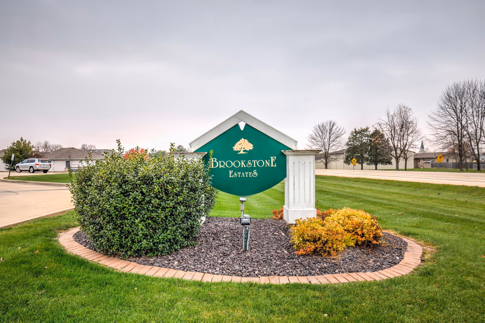 Outdoor view of a green and white sign for Brookstone Estates surrounded by bushes and landscaping with a road and houses in the background under a cloudy sky.