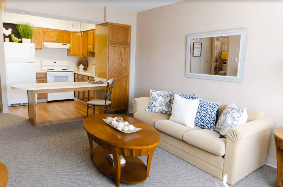 A cozy living area with a beige sofa adorned with patterned and plain cushions, a wooden oval coffee table with decorative items, and a kitchen in the background featuring wooden cabinets, a white refrigerator, stove, and a small breakfast bar with a chair.