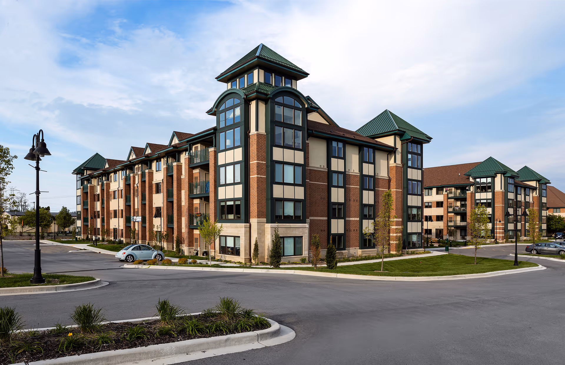 Exterior view of a multi-story senior living community building with brick and beige walls, green roofs, large windows, and landscaped surroundings including small trees and grass. There is a parking lot with a few cars and street lamps under a partly cloudy sky.