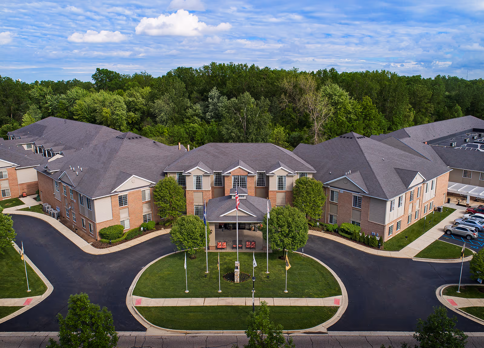 Aerial view of American House Park Place, a large senior living facility with multiple connected buildings surrounded by trees. The entrance features a covered drop-off area with flagpoles and a circular driveway. There are parked cars visible on the right side and well-maintained landscaping around the buildings.