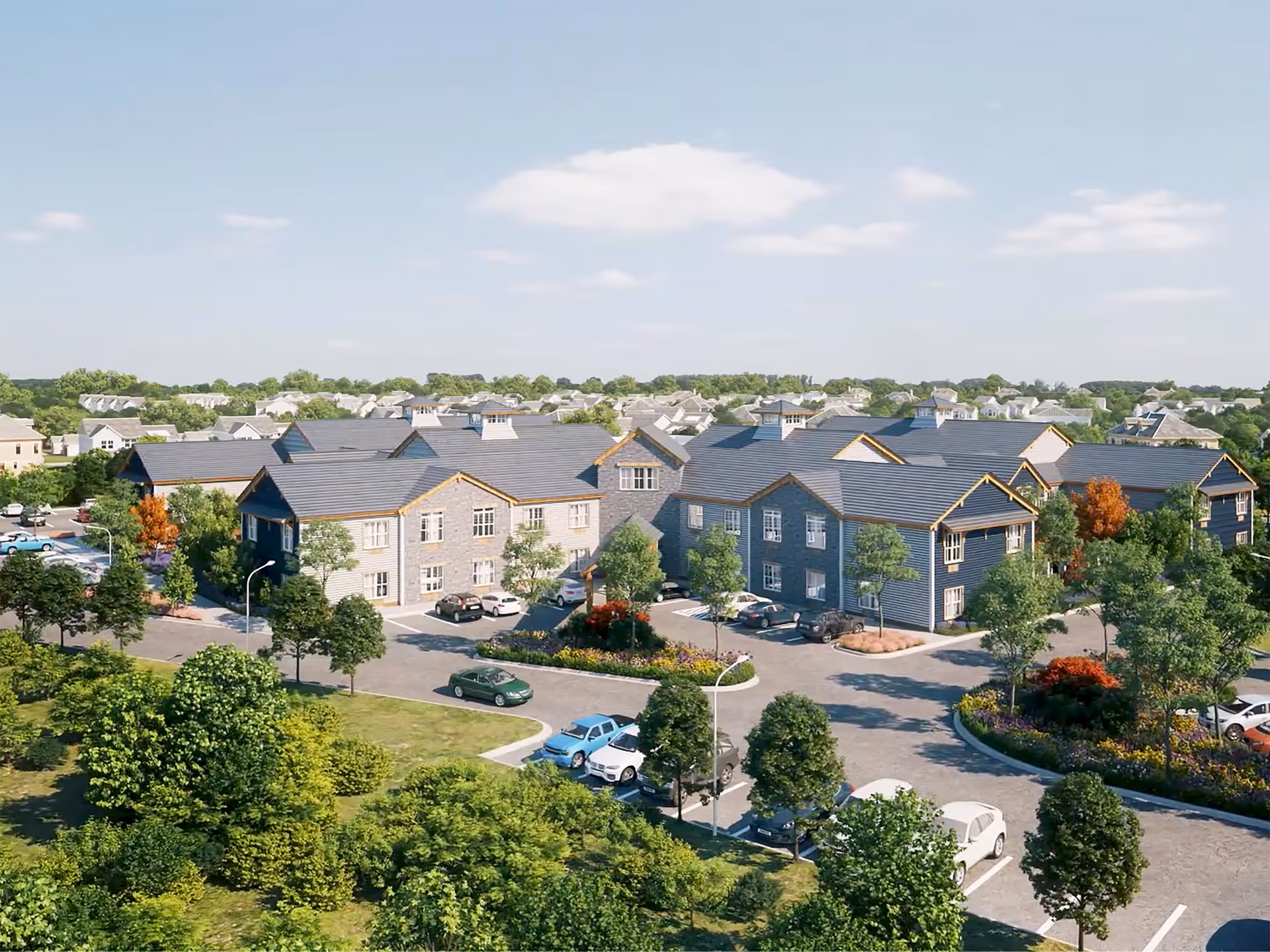 Aerial view of English Meadows Manassas Campus showing multiple two-story residential buildings with gray roofs and beige siding surrounded by trees, landscaped gardens, and parking areas with cars. The background includes a suburban neighborhood under a partly cloudy sky.