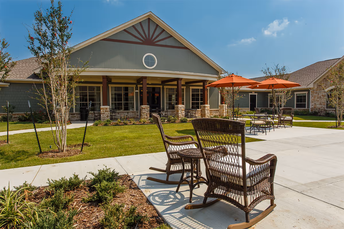 Outdoor courtyard and entrance of a single-story senior living building with wicker rocking chairs, patio tables and orange umbrellas on a sunny day.