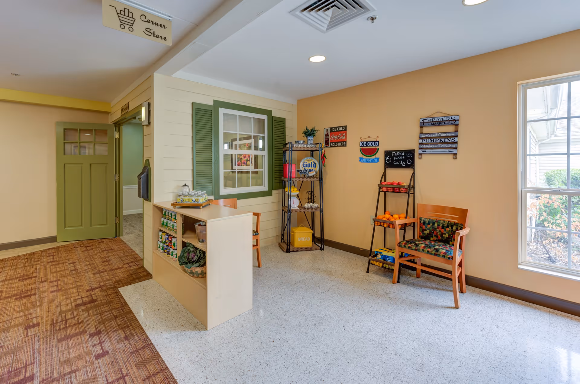 Interior view of a small corner store area inside a facility with a beige and green color scheme. There is a counter with shelves stocked with canned goods and other items, a green door, a window with green shutters, a chair with a colorful cushion, and a rack holding oranges and other products. Various vintage-style signs are displayed on the walls, and a window lets in natural light.