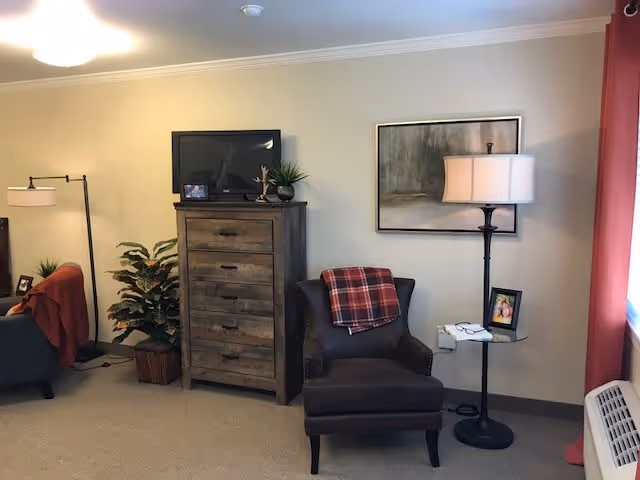A cozy living room area with a dark armchair draped with a red plaid blanket, a tall wooden dresser with a flat-screen TV on top, a floor lamp, a small round side table with a photo frame and glasses, a potted plant, and a framed abstract painting on the wall. The room has beige walls and carpeted floor with a window covered by red curtains.