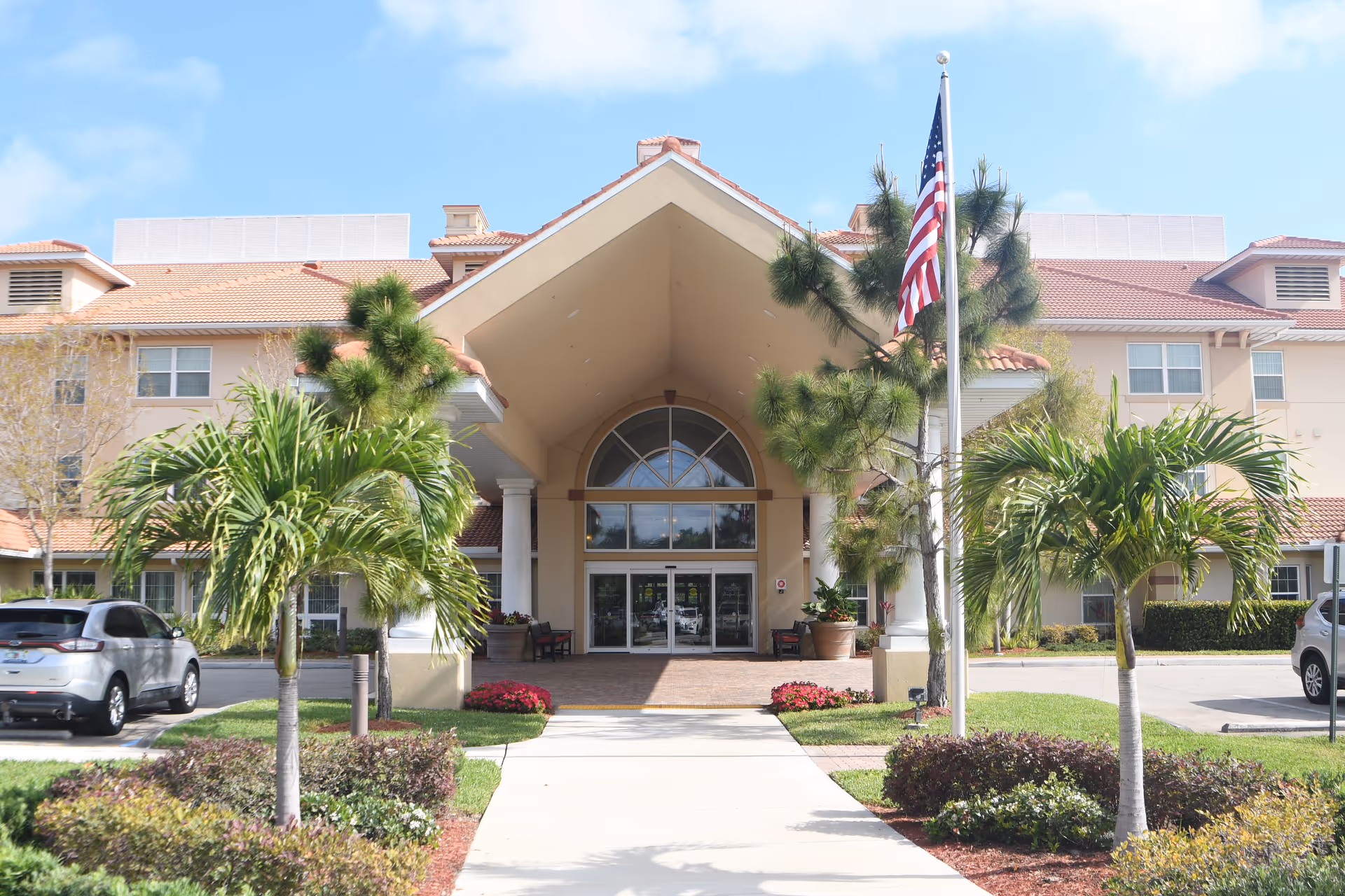 Front exterior view of Bradford Square Retirement Community building with a covered entrance, palm trees, an American flag on a flagpole, and parked cars on either side of the driveway.