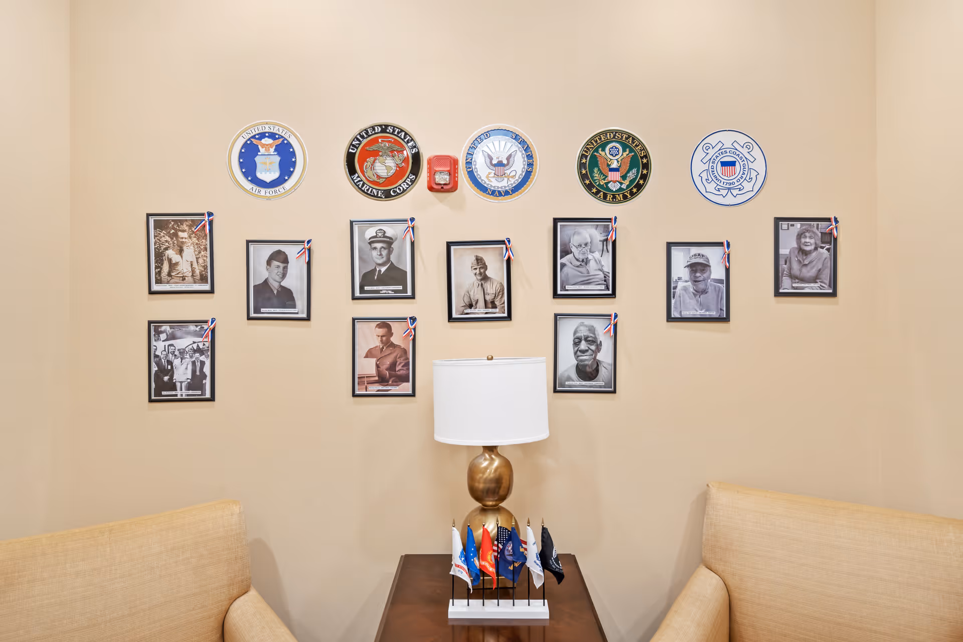 A beige wall decorated with framed black and white photographs of veterans, each adorned with a small red, white, and blue ribbon. Above the photos are circular emblems representing different branches of the United States military: Air Force, Marine Corps, Navy, Army, and Coast Guard. Below the photos is a wooden table with a gold lamp and a set of small flags representing various military branches. Two beige upholstered chairs flank the table on either side.