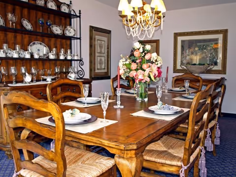 Wooden dining table set for six with crystal glassware, a floral centerpiece, chandelier overhead and a china cabinet in the background.