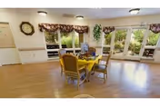 Bright dining area with a round table covered by a yellow tablecloth, chairs, a TV, wall clock, and large windows/doors overlooking greenery.