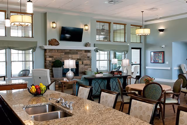 A spacious and well-lit common area in a senior living facility featuring a granite countertop with a sink and fruit bowl, several tables and chairs, a stone fireplace with a mounted flat-screen TV above it, decorative lamps, and large windows with blinds allowing natural light to fill the room.