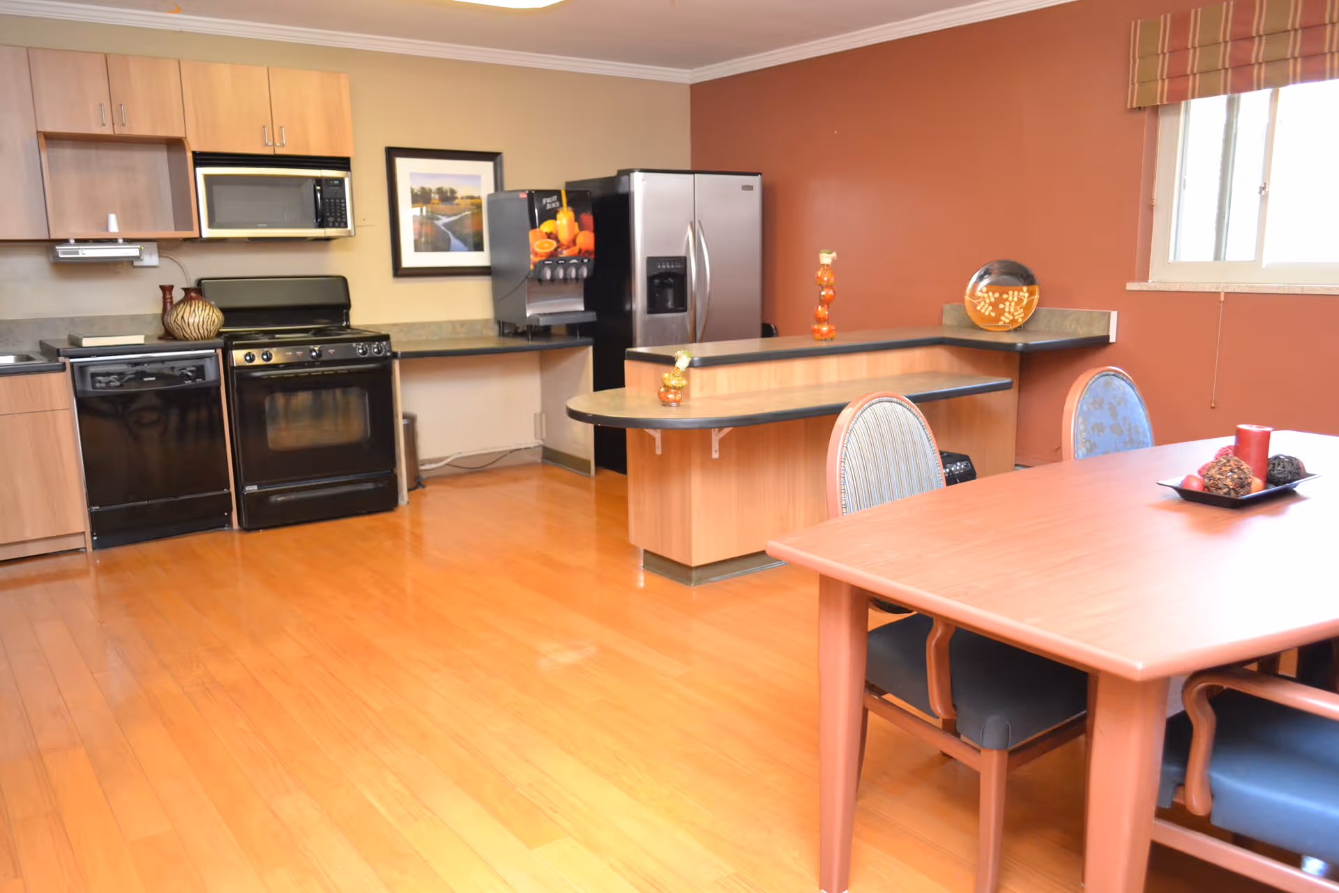 A kitchen and dining area with wooden floors and cabinetry. The kitchen features a black dishwasher, black oven with stove, microwave, and a stainless steel refrigerator. There is a counter with a beverage dispenser and decorative items. The dining area has a wooden table with four chairs and a window with a striped valance.