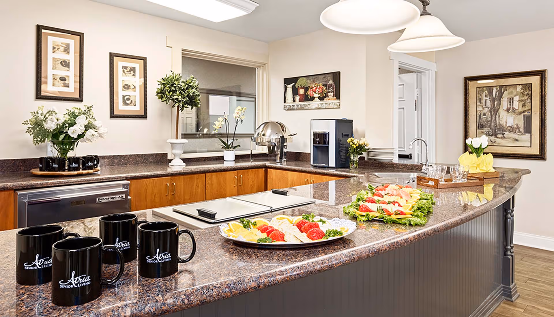 A clean and well-lit kitchen area with a curved granite countertop. On the counter, there are black mugs with 'Atria Senior Living' printed on them, plates of sliced fruits and crackers, a tray with glasses, and a coffee machine. The background features wooden cabinets, framed artwork on the walls, and decorative plants.