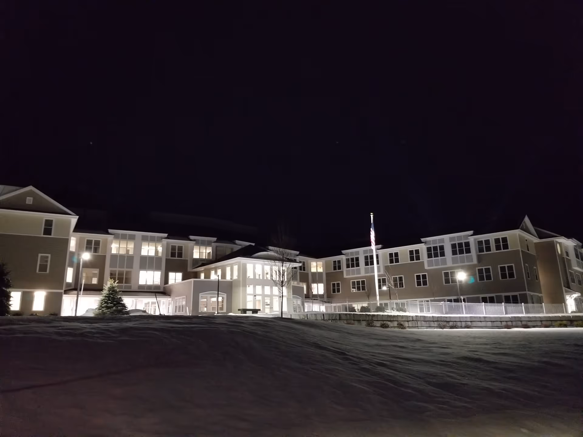 Nighttime exterior view of a large, well-lit senior living facility building with multiple windows, an American flag on a flagpole, and snow-covered ground in front.