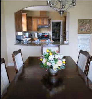 Dining room with a dark wooden table and six chairs, a floral centerpiece on the table, and a view into a kitchen with wooden cabinets and stainless steel appliances through an arched opening.